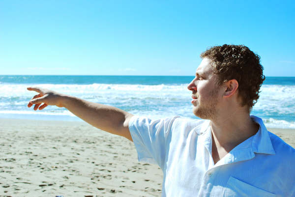 Man Points on the Beach: A man in a white shirt is standing on a beach pointing to the side with waves crashing in the background.
